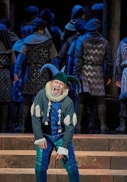 Błazen w zielono-niebieskim stroju i błazeńskiej czapce na scenie, za nim odwróceni tyłem do niego aktorzy, wszyscy w podobnych strojach i nakryciach głowy.