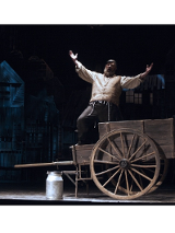 Mężczyzna ubrany w jasną szeroką koszulę, kamizelkę i czarne spodnie stoi na drewnianym wozie. Śpiewa wyciągając ręce przed siebie. Przed wozem stoi kanka na mleko.