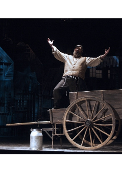 Mężczyzna ubrany w jasną szeroką koszulę, kamizelkę i czarne spodnie stoi na drewnianym wozie. Śpiewa wyciągając ręce przed siebie. Przed wozem stoi kanka na mleko.