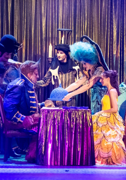 Przy stoliku siedzi kobieta w żółtej sukni, naprzeciw niej siedzi postawny mężczyzna w granatowym stroju, z rogami na głowie. Obok stołu stoją trzy postacie w strojach scenicznych, jedna z nich trzyma nad stolikiem półokrągłe naczynie.