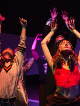 Kilka osób podczas występu. Na pierwszym planie dwie osoby, z podniesionymi rękoma i uniesionymi głowami. Ciemne tło.