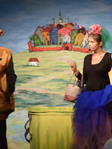 Mężczyzna ubrany w brązowy kostium, w niedużej odległości od niego kobieta trzymajaca kilka toreb, która patrzy na mężczyznę. W tle m.in. rysunek kilku domków otoczonych niewysokim murem.