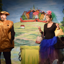 Mężczyzna ubrany w brązowy kostium, w niedużej odległości od niego kobieta trzymajaca kilka toreb, która patrzy na mężczyznę. W tle m.in. rysunek kilku domków otoczonych niewysokim murem.