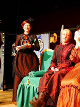 Scena ze spektaklu: cztery kobiety w strojach długich sukniach, dwie z nich siedzą na zielonej sofie, dwie stoją.