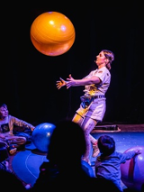 Kobieta na scenie podrzuca dużą pomarańczową piłkę. Widocznych jest kilka obserwujących ją osób. Ciemne tło.