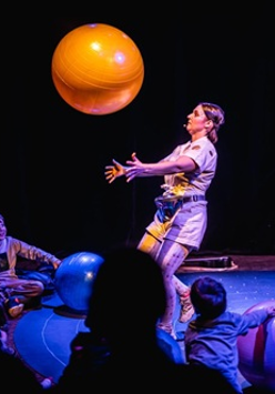 Kobieta na scenie podrzuca dużą pomarańczową piłkę. Widocznych jest kilka obserwujących ją osób. Ciemne tło.