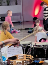 Na pierwszym planie kobieta grająca na perkusji odwrócona tyłem do obiektywu, na drugim planie kilkoro dzieci bawiących się na podłodze.