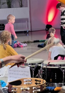 Na pierwszym planie kobieta grająca na perkusji odwrócona tyłem do obiektywu, na drugim planie kilkoro dzieci bawiących się na podłodze.