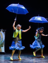 Dwie kobiety na scenie, każda z nich trzyma nad głową granatowy parasol.