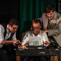 Trzech mężczyzn, jeden z nich siedzi przy stole i pisze coś na maszynie do pisania, Dwóch pozostałych stoi obok niego - jeden z nich trzyma ręce na barkach piszącego.