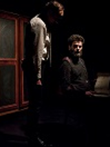 Ciemne zdjęcie: dwóch mężczyzn, jeden siedzi , drugi przy nim stoi. W tle drzwi szafy.