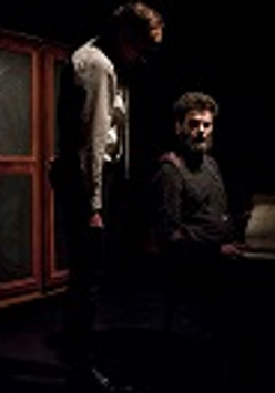 Ciemne zdjęcie: dwóch mężczyzn, jeden siedzi , drugi przy nim stoi. W tle drzwi szafy.