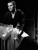 Czarno-białe zdjęcie, na którym jeden mężczyzna podtrzymuje drugiego mężczyznę. Ciemne tło, na drugim planie czterokołowy wózek kelnerski.