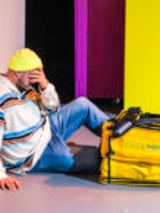 na zdjęciu mężczyzna siedzi pod ścianą, ręką zasłania twarz, przed nim na podłodze leży żółta torba termiczna do przewozu jedzenia