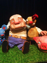 Lalka przedstawiająca starszego mężczyzny obejmującego myszkę, koguta i świnkę. Czarne tło.