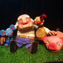 Lalka przedstawiająca starszego mężczyzny obejmującego myszkę, koguta i świnkę. Czarne tło.