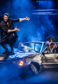 Zdjęcie przedstawiające trzy osoby wysiadające ze zniszczonego samochodu oraz jednego mężczyznę stojącego na masce samochodu.