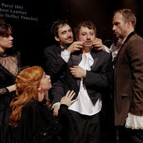 Dwóch mężczyzn soi blisko trzeciego mężczyzny, jeden z nich dotyka jego twarzy. Dwie kobiety przygladają się mężczyznom.