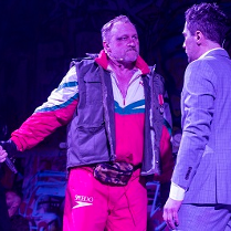 Dwóch mężczyzn stojących naprzeciwko siebie, jeden eleganco ubrany w garnitur, drugi w sportowym dresie i ortalionowej kamizelce.