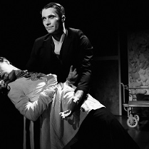 Czarno-białe zdjęcie, na którym jeden mężczyzna podtrzymuje drugiego mężczyznę. Ciemne tło, na drugim planie czterokołowy wózek kelnerski.