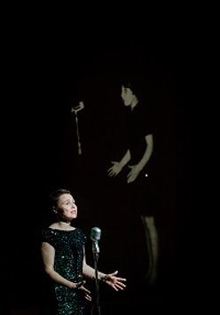 Kobieta w czarnej sukience stoi przed mikrofonem, za nią zdjęcie innej kobiety stojącej przed mikrofonem na czarnym tle.