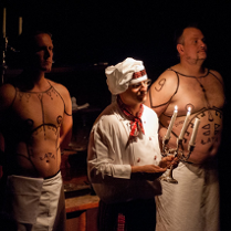 Na zdjęciu trzech mężczyzn: jeden, w środku, ubrany w biały uniform z biała kucharską czapką na głowie, trzyma w ręce świecznik, dwóch pozostałych z nagimi wytatułowanymi torsami za nim.