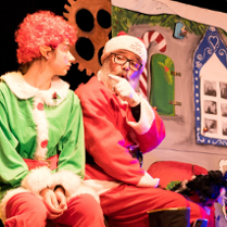 Zdjęcie ze spektaklu: obok siebie siedzą świety Mikołaj i elf, Mikołaj wskazuje palcem na elfa, coś mu tłumaczy. W tle elementy świątecznej scenografii.
