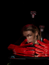 Na ciemnym tle twarz kobiety, przed nią duże czerwone rękawiczki.