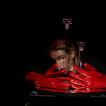 Na ciemnym tle twarz kobiety, przed nią duże czerwone rękawiczki.