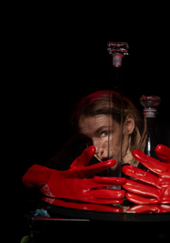 Na ciemnym tle twarz kobiety, przed nią duże czerwone rękawiczki.