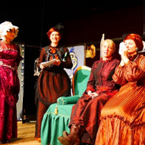 Scena ze spektaklu: cztery kobiety w strojach długich sukniach, dwie z nich siedzą na zielonej sofie, dwie stoją.