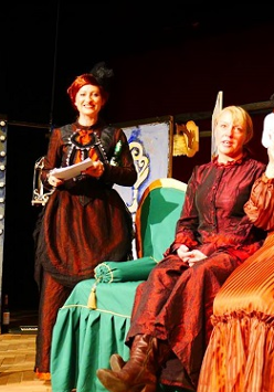 Scena ze spektaklu: cztery kobiety w strojach długich sukniach, dwie z nich siedzą na zielonej sofie, dwie stoją.