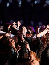 Pośrodku sceny stoi mężczyzna, wznosi ręce w górę, śmieje się, ciasno otacza go tłum ludzi, którzy starają się go dotknąć, śmieją się