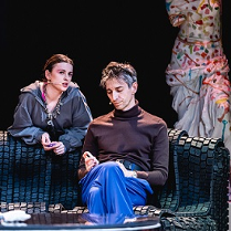 Mężczyzna siedzi na kanapie, za kanapą kobieta, która jest o nią oparta i skierowana w stronę mężczyzny. W tle fragment manekinu, na którym wisi kolorowa suknia.