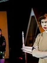 Na pierwszym planie kobieta z założonymi rękami patrzy przed siebie, na drugim planie druga kobieta na tle otwartych drzwi.