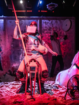 Mężczyzna z zakrytą twarzą siedzi na drewnianym siedzisku przypominającym siodło, trzyma w ręce długi kij. Po prawej stronie figura konia do połowy zakopana w piasku.