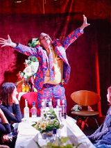 Przy stole siedzi kilka osób, u szczytu stołu stoi mężczyzna w kolorowym garniturze z szeroko rozłożonymi rękoma. Na stole kilka butelek alkoholu.