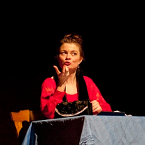 Dwie kobiety, jedna siedzi przy stole i gestykuluje, druga stojąc z założonymi rękoma patrzy w jej kierunku. Ciemne tło.