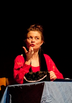 Dwie kobiety, jedna siedzi przy stole i gestykuluje, druga stojąc z założonymi rękoma patrzy w jej kierunku. Ciemne tło.