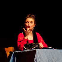 Dwie kobiety, jedna siedzi przy stole i gestykuluje, druga stojąc z założonymi rękoma patrzy w jej kierunku. Ciemne tło.