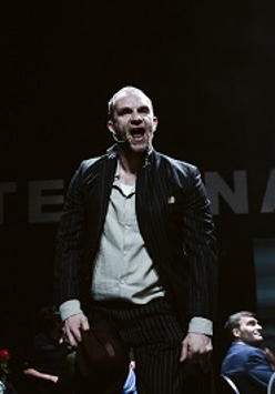 Mężczyzna, który stoi z otwartymi ustami, wokół niego siedzące na krzesłach osoby. Ciemne tło.