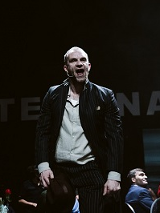 Mężczyzna, który stoi z otwartymi ustami, wokół niego siedzące na krzesłach osoby. Ciemne tło.