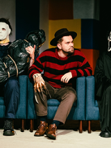 Trzy siedzące na fotelach postacie: w środku mężczyzna patrzący w bok, z jego obu stron postacie w maskach na twarzach.