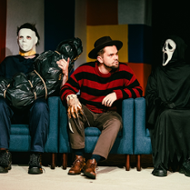 Trzy siedzące na fotelach postacie: w środku mężczyzna patrzący w bok, z jego obu stron postacie w maskach na twarzach.