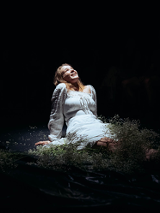 Na czarnym tle zdjęcie młodej, uśmiechniętej dziewczyny w białej sukni. Dziewczyna siedzi oparta rękoma o podłogę, patrzy ku górze.