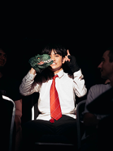 Na ciemnym tle mężczyzna w białej koszuli i czerwonym krawacie siedzi na fotelu, ma uniesione ręce.