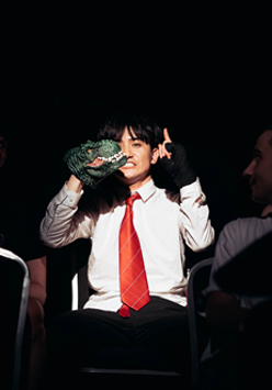 Na ciemnym tle mężczyzna w białej koszuli i czerwonym krawacie siedzi na fotelu, ma uniesione ręce.