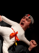 Scena z przedstawienia: mężczyzna trzymający lalkę ubraną w biały, sportowy strój, w miejscu głowy lalki widać głowę aktora. Poza lalki to poza karate z jedną nogą uniesioną do góry.