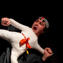 Scena z przedstawienia: mężczyzna trzymający lalkę ubraną w biały, sportowy strój, w miejscu głowy lalki widać głowę aktora. Poza lalki to poza karate z jedną nogą uniesioną do góry.