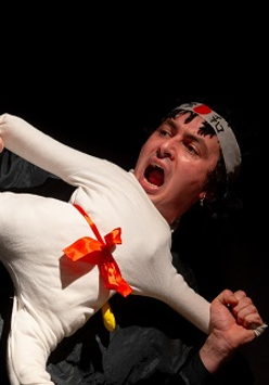 Scena z przedstawienia: mężczyzna trzymający lalkę ubraną w biały, sportowy strój, w miejscu głowy lalki widać głowę aktora. Poza lalki to poza karate z jedną nogą uniesioną do góry.